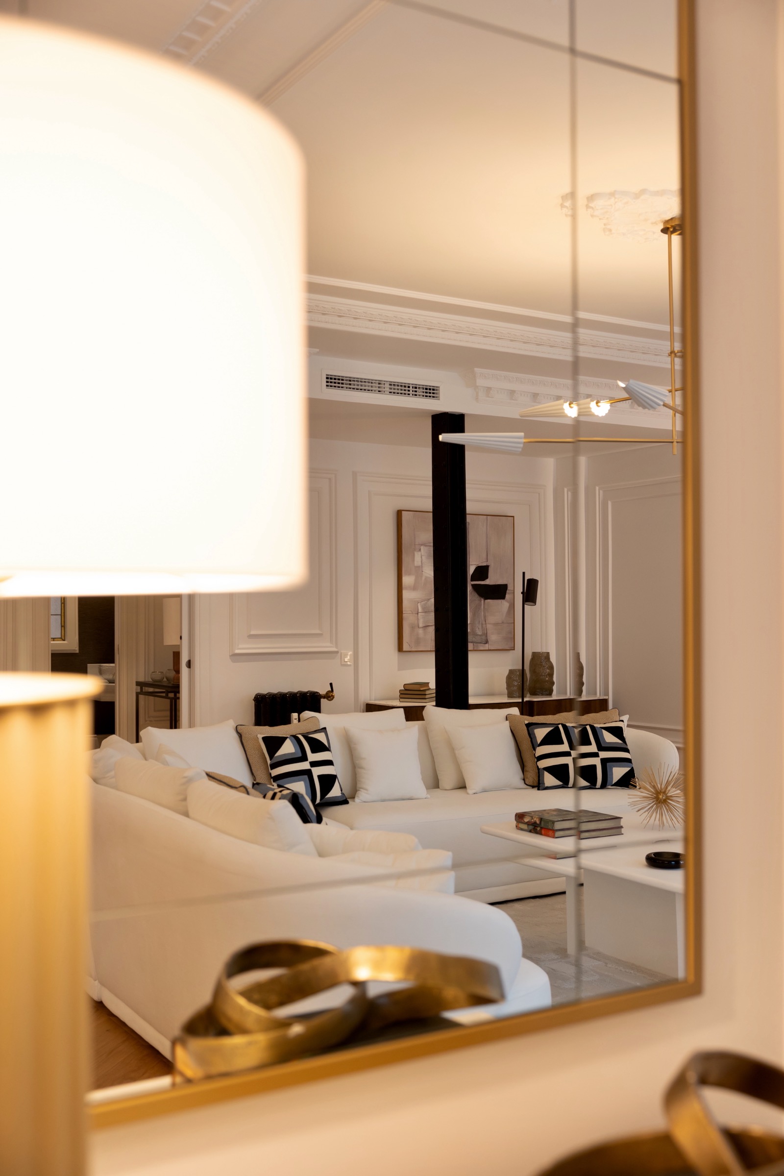 Elegante sala de estar con sofá blanco, cojines decorativos en blanco y negro, paredes blancas con molduras, lámpara de techo dorada y espejo que refleja la habitación. Decoración en tonos neutros con acentos dorados.