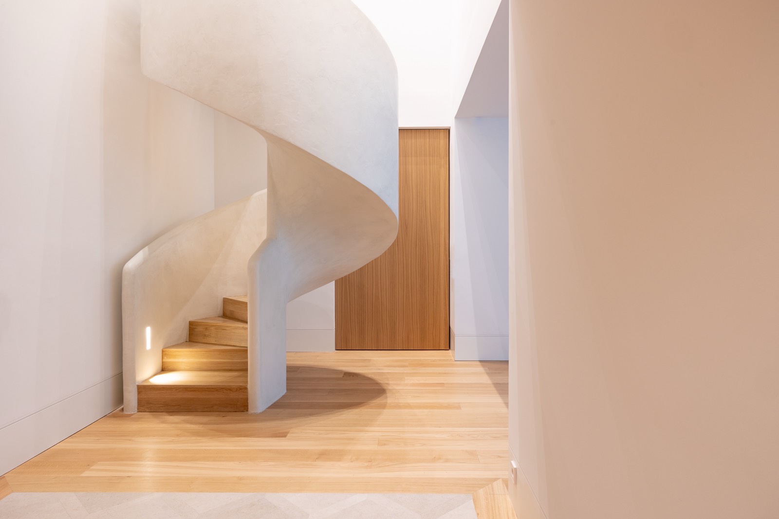 Escalera de caracol de madera con diseño curvo y paredes blancas, con iluminación suave y puerta de madera en tonos cálidos