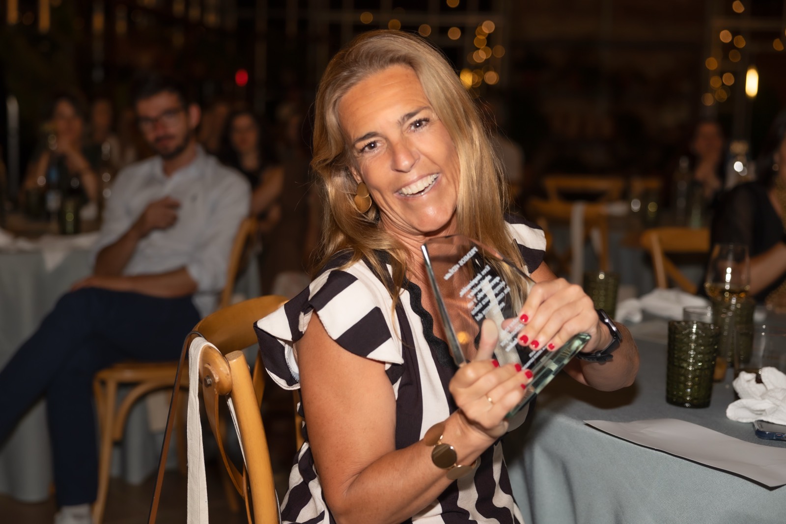 Mujer rubia con vestido blanco y negro sonriendo mientras sostiene una copa de vidrio en un evento social con iluminación tenue
